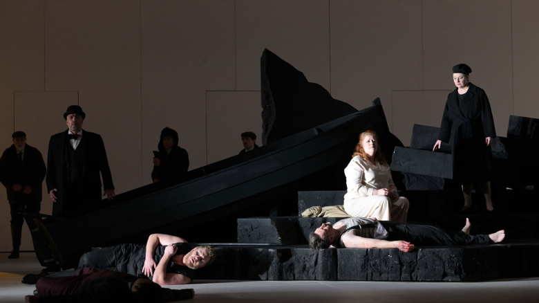 Andreas Bauer Kanabas (König Marke; mit Hut) sowie liegend Nicholas Brownlee (Kurwenal; mit ärmelloser Weste) und Taehan Kim (Melot; in rotem Anzug) sowie Marco Jentzsch (Tristan; liegend), Miina-Liisa Värelä (Isolde; sitzend) und Claudia Mahnke (Brangäne